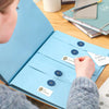 A person is opening a blue accordion document organizer file folder to view passports and birth certificates on a wooden tabl