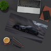 A large gaming mouse pad featuring a black JDM car design on a gray road surface, next to a laptop and writing tools.