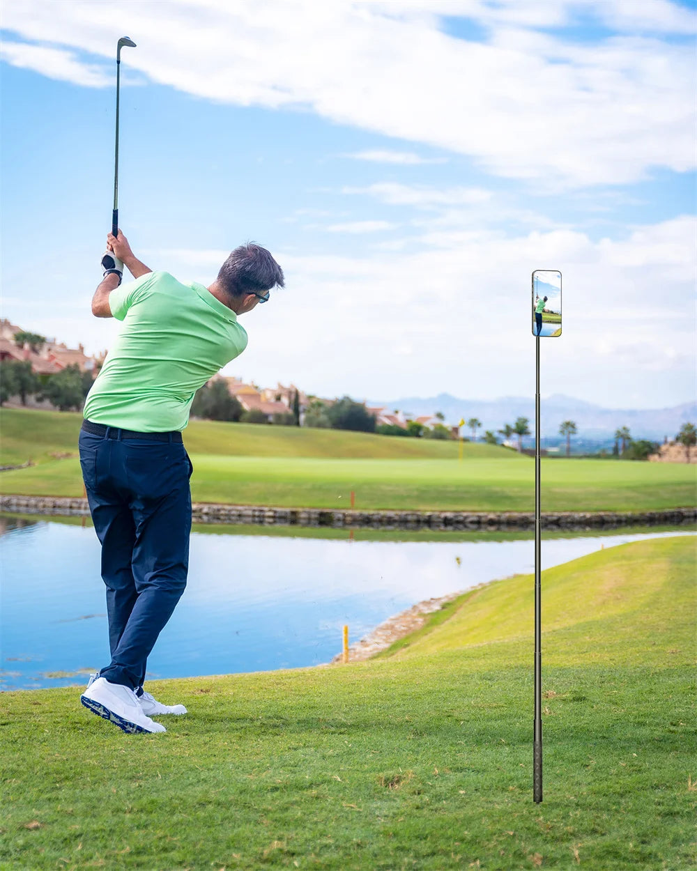 A man in a green shirt is swinging a golf club while a magnetic monopod records the shot.