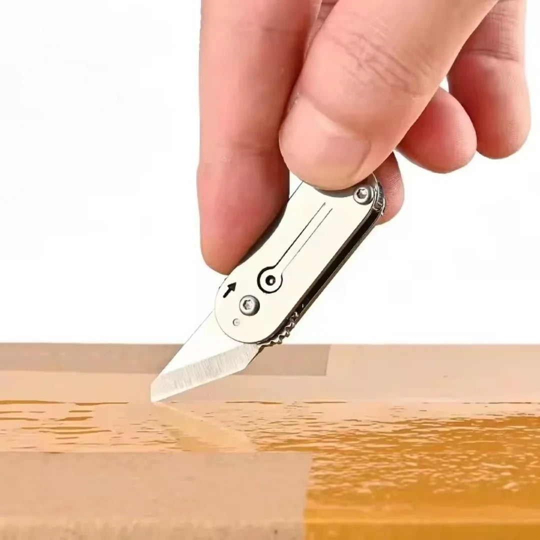 A hand holds a compact stainless steel pocket knife with a keychain, showing the blade tip resting on a wooden surface.