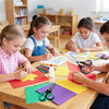 A boy and a girl are using a multipurpose stainless steel scissors set to cut colored paper at a wooden table.