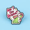 An enamel pin featuring a green frog character with a pink heart and text that reads Am I the Drama