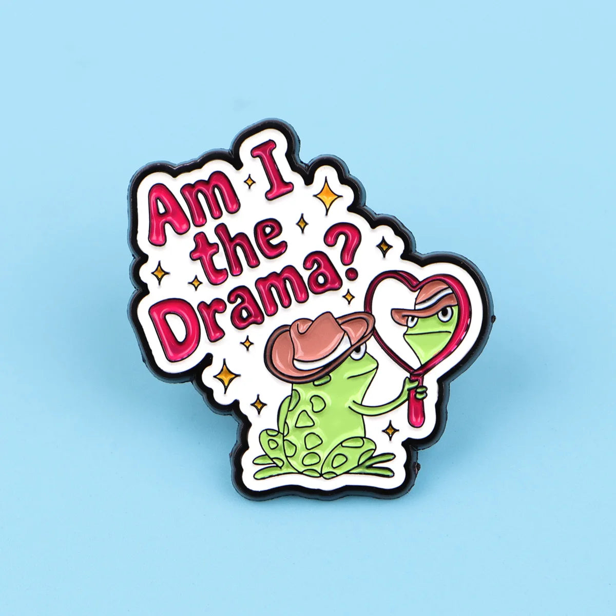 An enamel pin featuring a green frog character with a pink heart and text that reads Am I the Drama