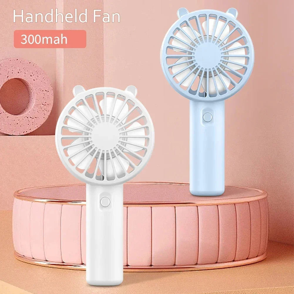 A light blue handheld fan with a white grille is displayed on a pink circular stand.