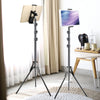 A black adjustable tripod mount is shown with a tablet attached, next to another similar mount and a wooden table.