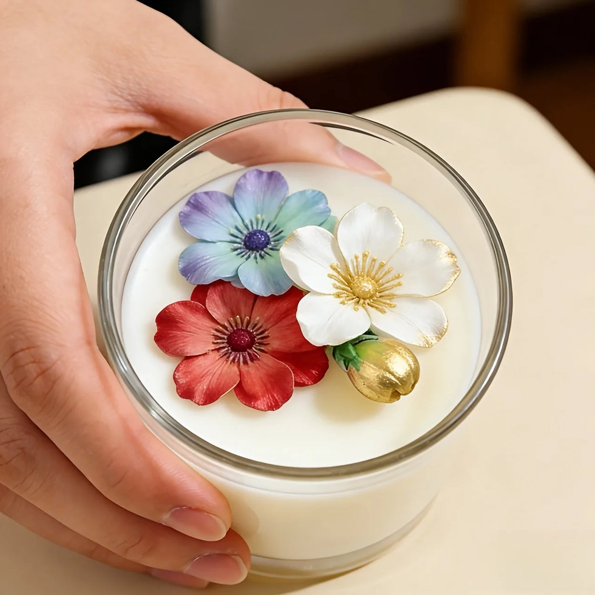 A hand holds a glass of white candle wax decorated with colorful floral wax flowers.