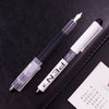 A black leather pouch holds a clear fine point fountain pen and a white pen, with a roll of paper visible below.