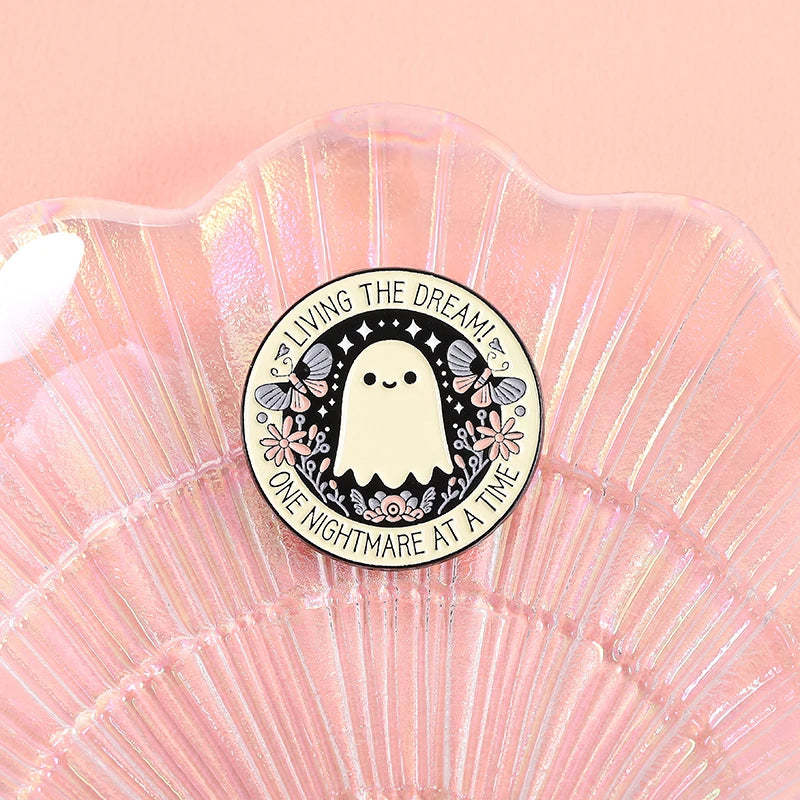 A round enamel pin featuring a cute white ghost with pink floral accents on a translucent pink shell background.
