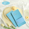 A blue slim weekly planner journal is displayed on a light background with yellow and white accents and yellow donuts.