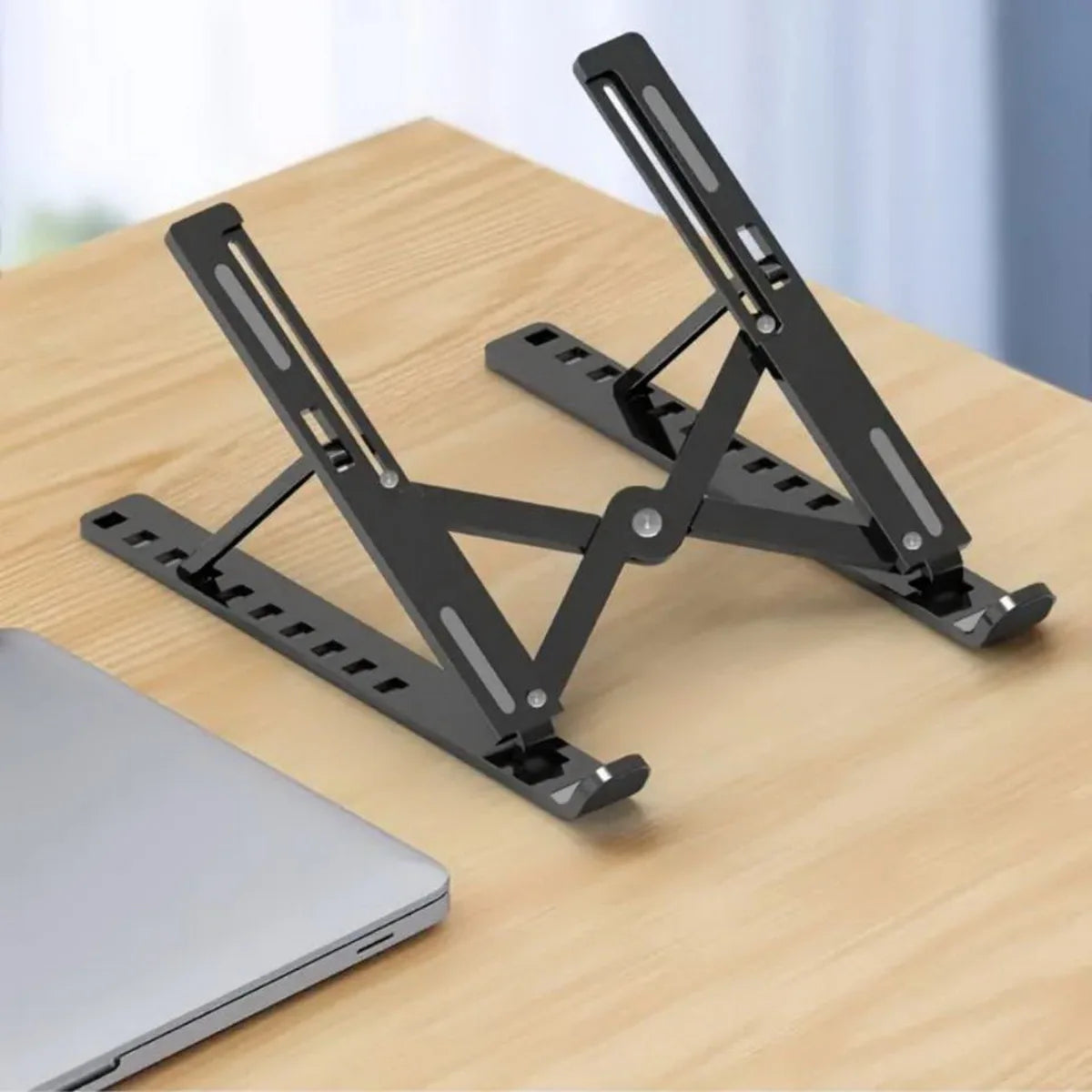 A black portable folding laptop stand is shown resting on a wooden surface next to a silver laptop.