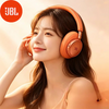 A woman wearing orange JBL headphones, smiling and touching her ear, in soft lighting.
