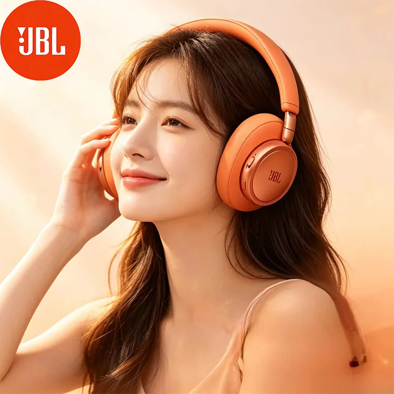 A woman wearing orange JBL headphones, smiling and touching her ear, in soft lighting.