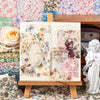An open scrapbook shows floral paper designs and decorative elements on a wooden stand.
