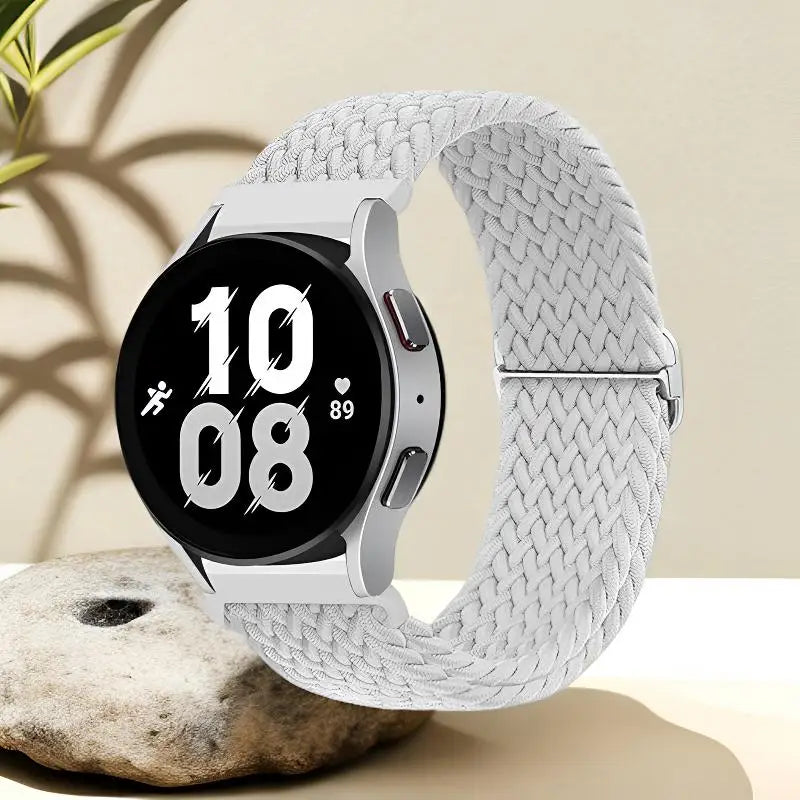 A silver nylon watch strap is shown coiled around a smartwatch, resting on a light-colored stone.