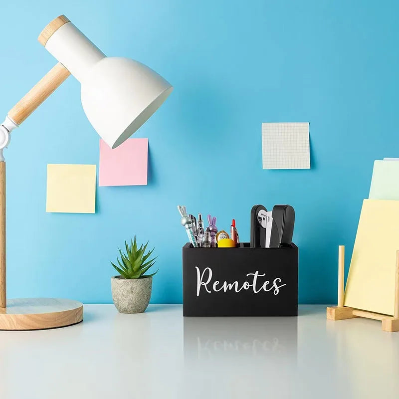 A black storage box with dividers for remote controls sits on a light blue desk next to a wooden desk lamp.