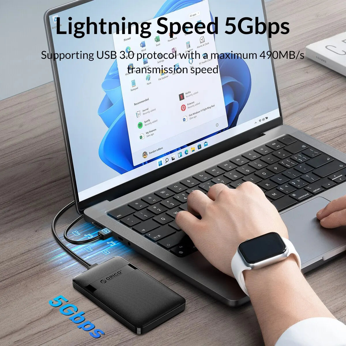 A hand is using a portable 5Gbps USB drive enclosure connected to a laptop, demonstrating high-speed data transfer.