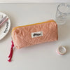 A pink checkered pencil case with a yellow trim rests on a white surface next to a glass and a silver ring.