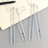 Tres reglas plegables de acrílico transparente con pompones blancos decorativos, exhibidas sobre un cuaderno rayado.