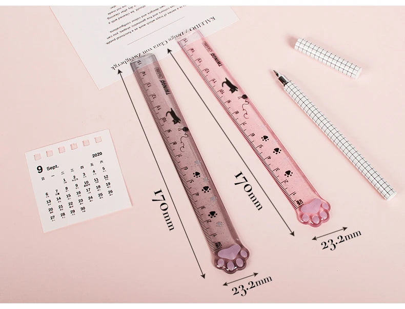 A pink plastic measuring tool shaped like a cat paw is shown next to a calendar and a ruler.