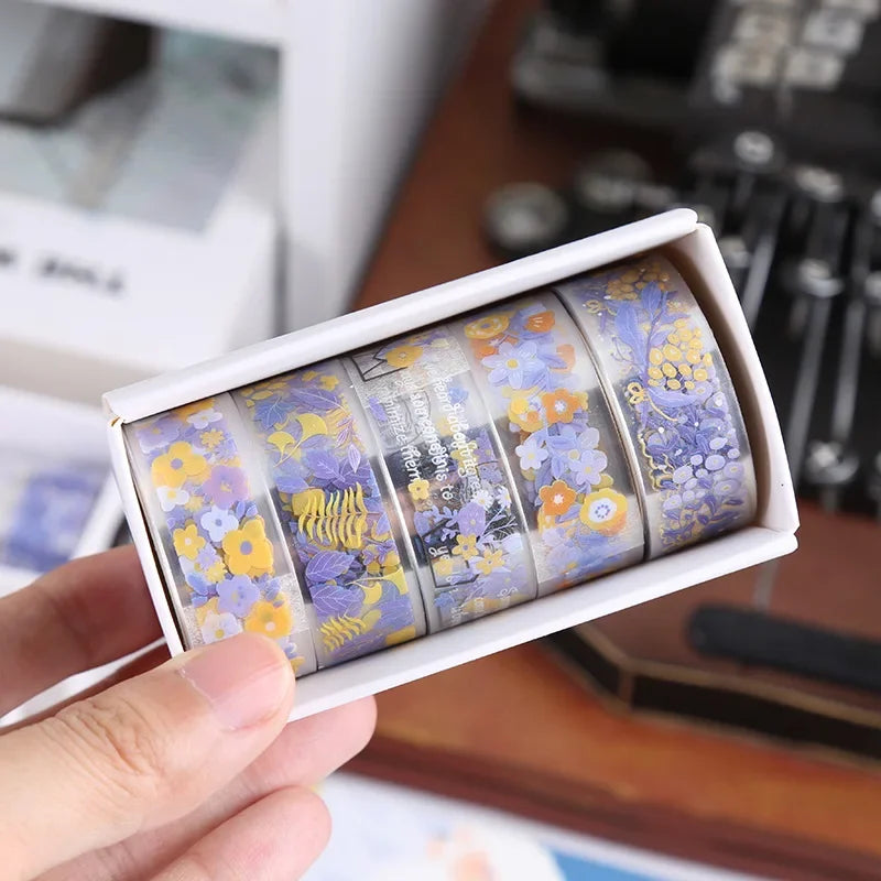 A set of decorative transparent washi tape rolls featuring floral patterns in purple, yellow, and white.