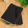 A stack of black A6 blank sketchbooks with a black pen resting on top, placed on a light wooden surface.