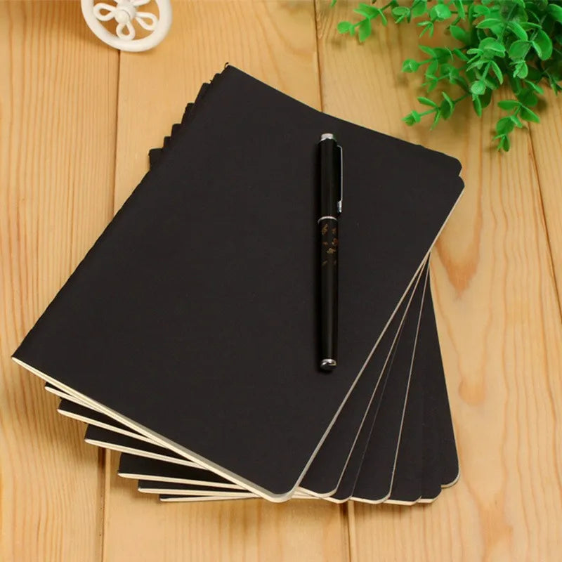 A stack of black A6 blank sketchbooks with a black pen resting on top, placed on a light wooden surface.
