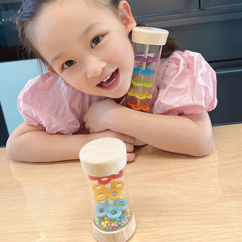 A young girl is holding a wooden rain sound tube musical instrument filled with colorful beads and other objects.