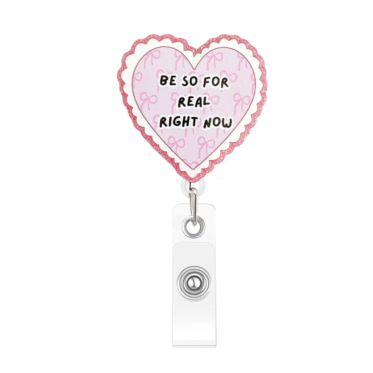A pink heart-shaped badge reel with a scalloped edge and pink ribbon details, featuring the text BE SO FOR REAL RIGHT NOW.