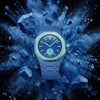 A close-up of a blue ceramic watch with a matching strap, surrounded by an explosion of blue dust.