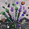 A collection of purple and green bead pens with witch charms laid out on a dark gray surface.