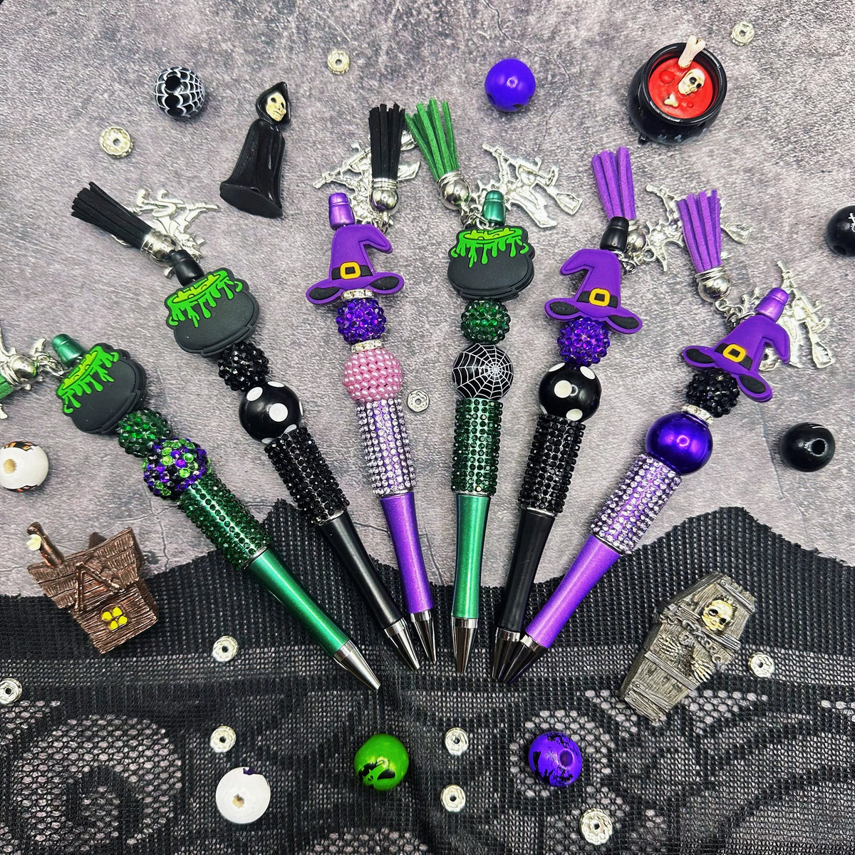 A collection of purple and green bead pens with witch charms laid out on a dark gray surface.