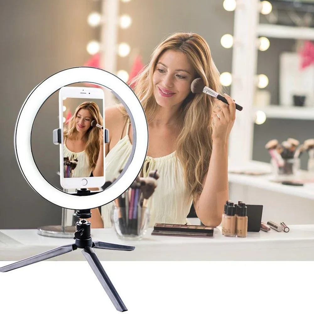 A woman uses an LED ring light with a tripod stand to film herself for makeup or streaming.