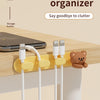 A yellow cable organizer with multiple white cables is shown on a wooden desk next to a laptop and a teddy bear.