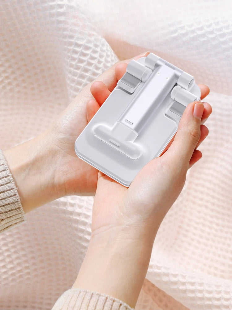 A person is holding a white foldable adjustable phone holder stand against a pink knitted background.