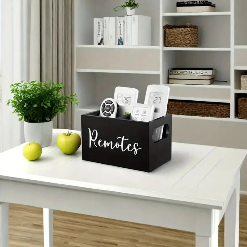 A black storage box labeled Remotes holding various remote controls and small electronics on a white table.