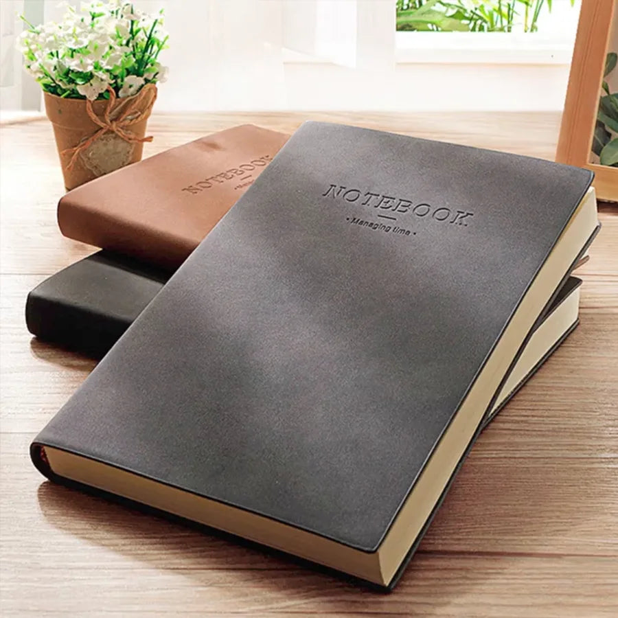 A dark gray notebook lying on a wooden surface next to a brown notebook and some green plants.