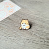 Un pequeño broche de esmalte redondo con forma de un lindo gato naranja y blanco sentado sobre una superficie de madera.