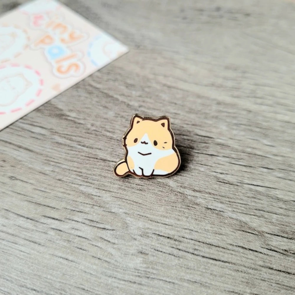 Un pequeño broche de esmalte redondo con forma de un lindo gato naranja y blanco sentado sobre una superficie de madera.