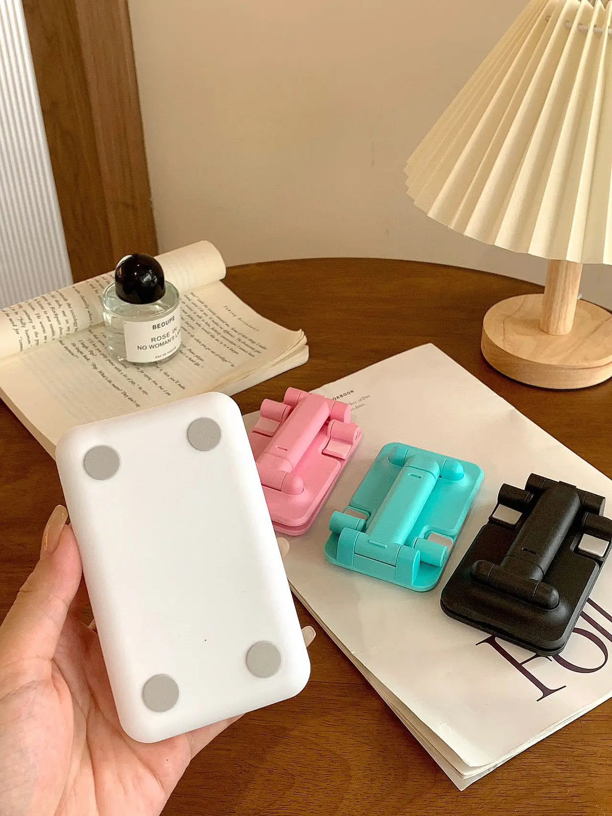 A hand holds a white adjustable foldable phone and tablet stand next to a stack of papers on a wooden table.