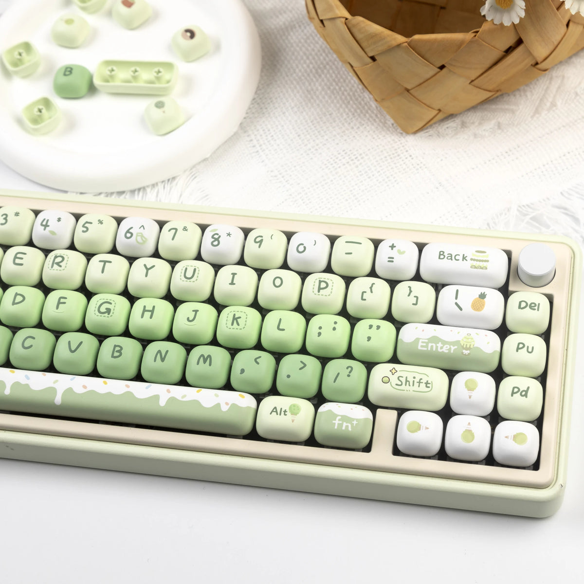 A close-up of a light green mechanical keyboard featuring colorful PBT keycaps and an apple-shaped keycap, next to a woven ba