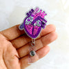 A hand holds a purple anatomical diagram badge reel featuring a heart illustration.