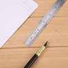A black pen rests on a wooden surface next to a stainless steel measuring ruler and a sheet of paper.