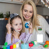 Eine blonde Frau hält die Hand eines jungen Mädchens, während sie bunte Glitzerartikel auf einem Tisch anschauen.