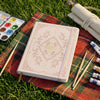 A white A5 dot grid notebook with a gold foil botanical cover rests on a plaid fabric surrounded by art supplies.