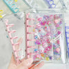 A hand holds a clear double-button binder filled with colorful stickers and small decorative items.