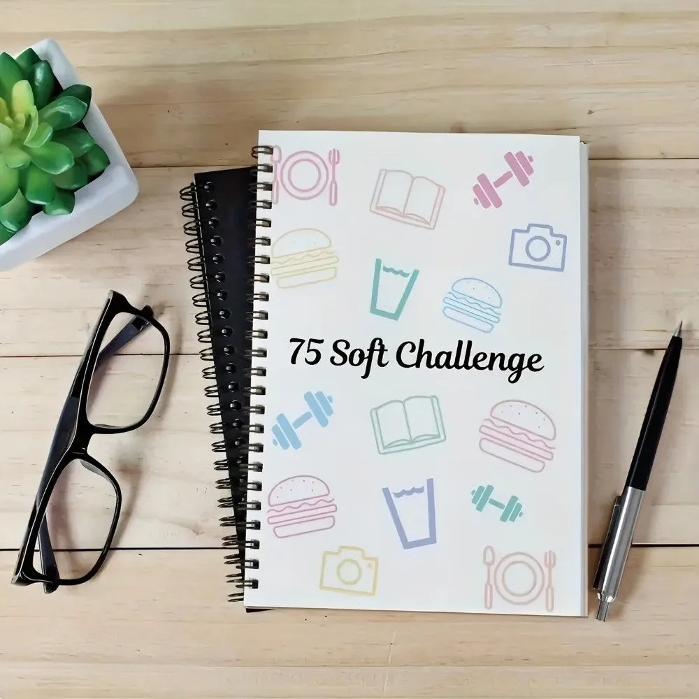 A spiral-bound notebook titled 75 Soft Challenge featuring various icons like dumbbells, books, and food items on a light bac