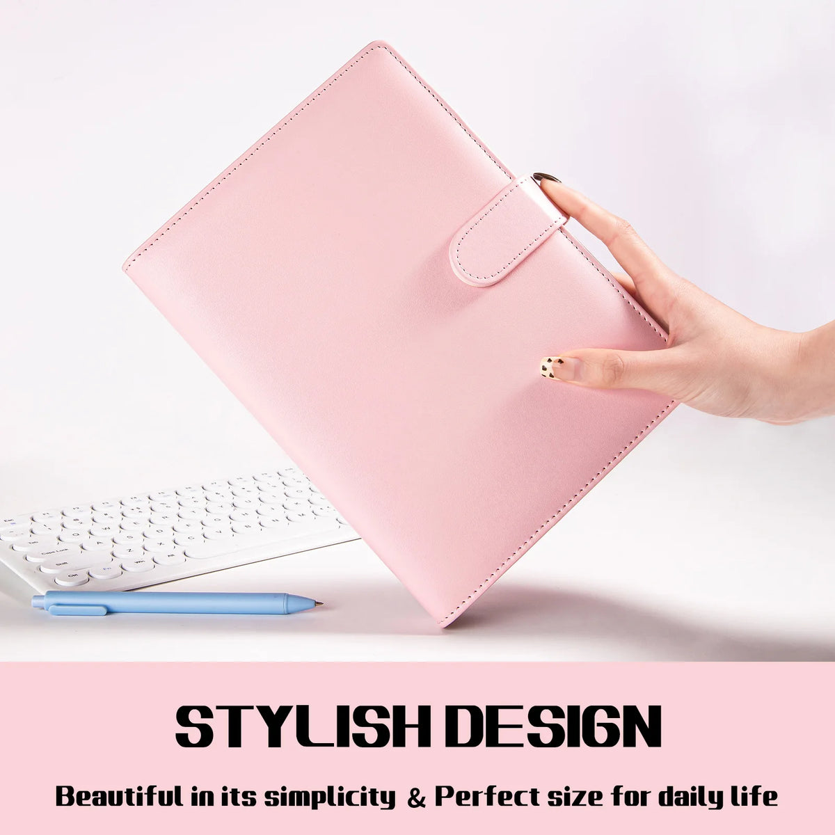 A pink A5 PU leather binder notebook with a cover flap is held by a hand next to a white keyboard and blue pen.