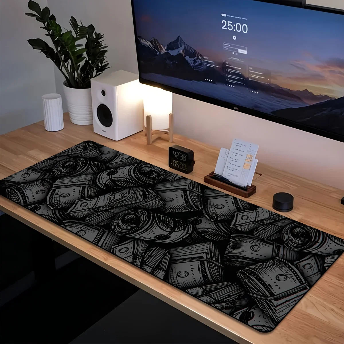 A large black and white desk mat with a pattern of stacked money is placed on a wooden desk in front of a computer monitor.