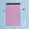 A pink lined writing pad with various fruit-themed stickers and a small orange tag on a light blue background.