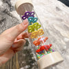 A hand holds a wooden rain sound tube musical instrument with colorful plastic flowers inside.